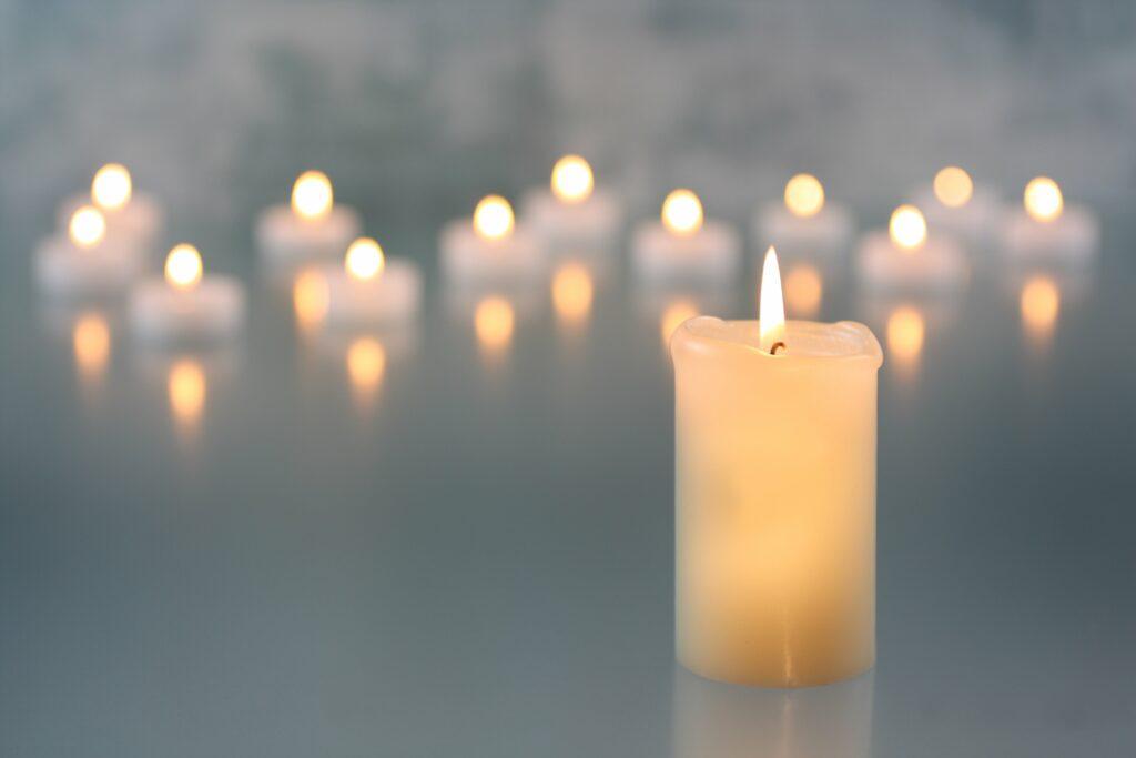 Ein Bild mit mehreren Kerzen, von denen eine im Vordergrund steht und brennt. Die anderen Kerzen im Hintergrund sind unscharf und verschwommen, was eine ruhige und besinnliche Atmosphäre schafft.