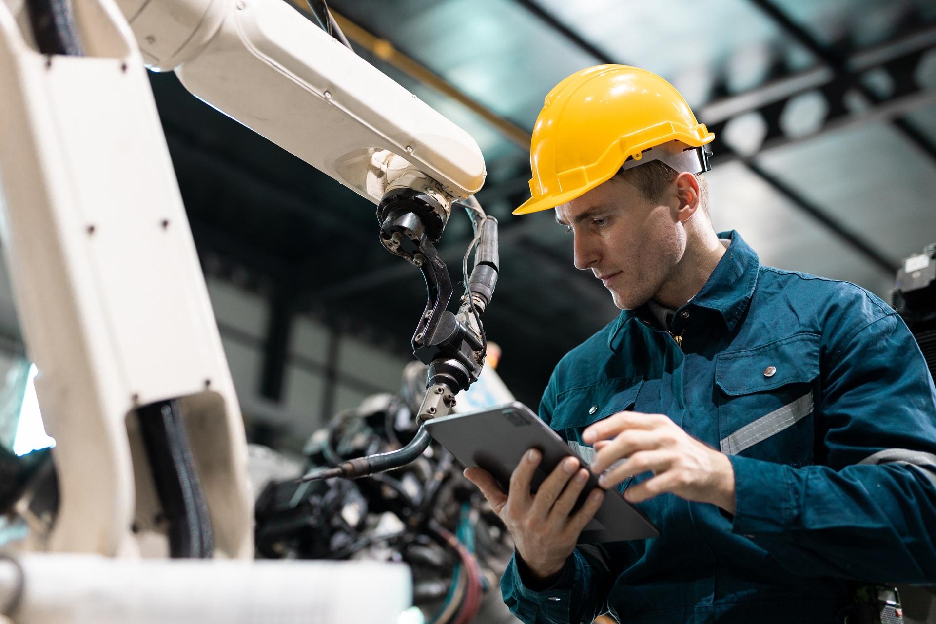 Ingenieur mit gelbem Helm arbeitet mit Tablet neben Roboterarm | E-Learning Anbieter